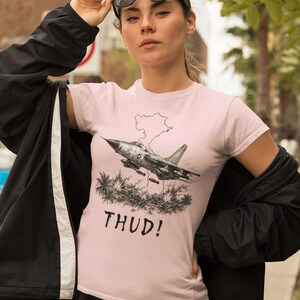 May include: A light pink t-shirt featuring a black and white graphic of a jet plane with a map outline and palm trees. The word "THUD!" is printed below the graphic. The person is wearing black sunglasses and a black jacket.