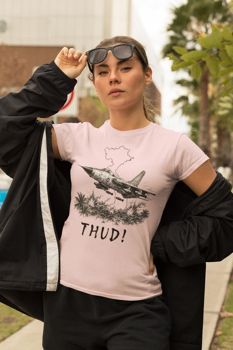 May include: A light pink t-shirt featuring a black and white graphic of a jet plane with a map outline and palm trees. The word "THUD!" is printed below the graphic. The person is wearing black sunglasses and a black jacket.