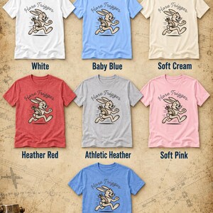 Puede incluir: Una colecci&oacute;n de siete camisetas de manga corta en blanco, azul beb&eacute;, crema suave, rojo jaspeado, gris jaspeado atl&eacute;tico, rosa suave y azul columbia jaspeado. Cada camiseta presenta un conejo corriendo con una granada y el texto Hare Trigger.