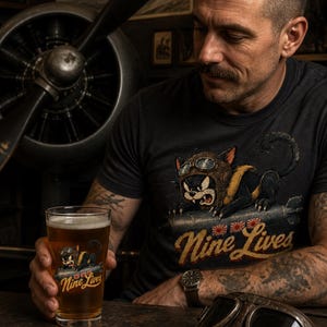 Op de afbeelding: Een zwart t-shirt en een helder glas, beide voorzien van een afbeelding van een zwarte kat met een vliegersbril op een bom, met de tekst "Nine Lives" in een geel lettertype boven drie kleine rood-witte rijzende zon-iconen.