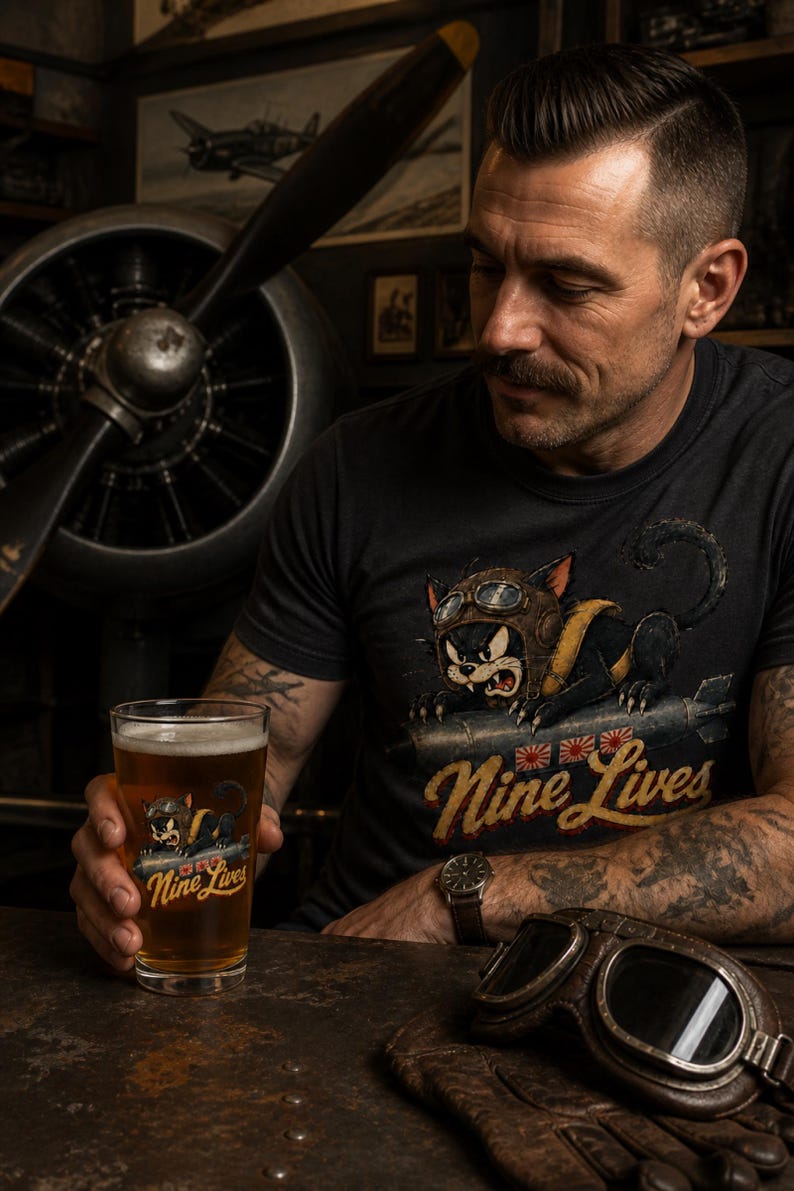 Op de afbeelding: Een zwart t-shirt en een helder glas, beide voorzien van een afbeelding van een zwarte kat met een vliegersbril op een bom, met de tekst "Nine Lives" in een geel lettertype boven drie kleine rood-witte rijzende zon-iconen.
