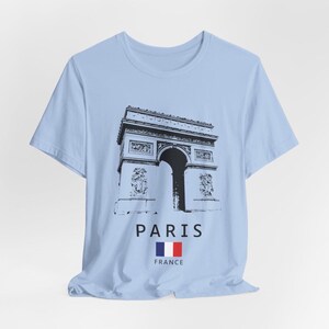 Puede incluir: Camiseta azul claro con una ilustraci&oacute;n en blanco y negro del Arco del Triunfo de Par&iacute;s. Debajo de la imagen, se lee la palabra "PARIS", junto con la bandera francesa y la palabra "FRANCE".