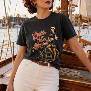 Op de afbeelding: Een antracietgrijs t-shirt met korte mouwen en een grafische print van een pin-up zeeman op een touwladder met een anker. De tekst luidt Permission to Come Aboard? in een rood lettertype. Het shirt heeft een ronde hals en een relaxte pasvorm.