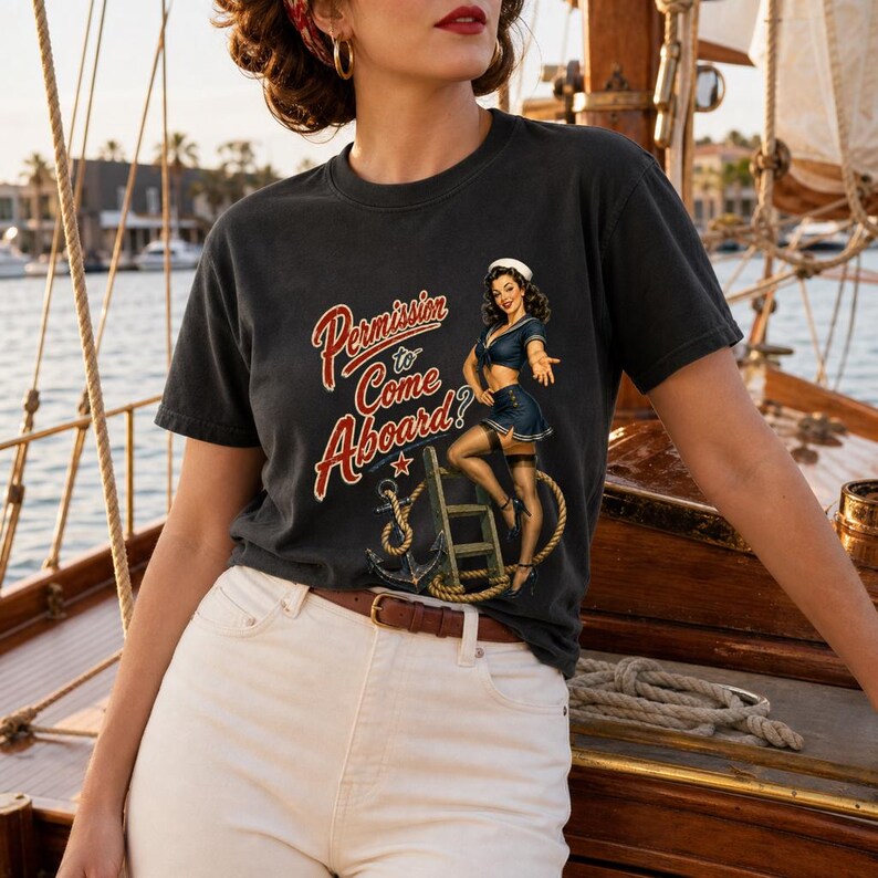 Op de afbeelding: Een antracietgrijs t-shirt met korte mouwen en een grafische print van een pin-up zeeman op een touwladder met een anker. De tekst luidt Permission to Come Aboard? in een rood lettertype. Het shirt heeft een ronde hals en een relaxte pasvorm.