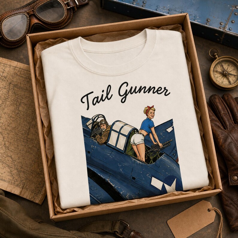 May include: A white t-shirt featuring a graphic print of a blue military aircraft with a pilot and a pin-up style figure in the tail gunner position. The text "Tail Gunner" is printed in black script above the illustration of the airplane.