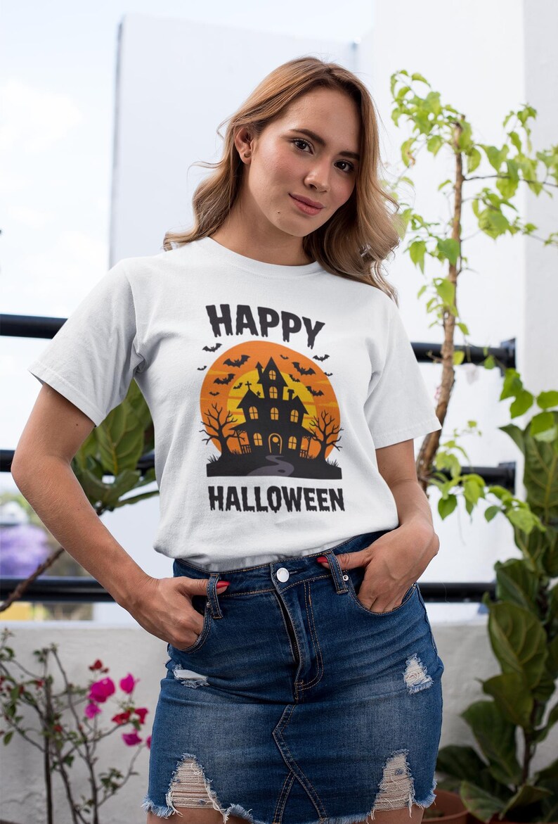 May include: White t-shirt with a Halloween design. The graphic features a haunted house silhouette against an orange sunset, with bats and the words "HAPPY HALLOWEEN". The shirt is paired with a denim skirt.