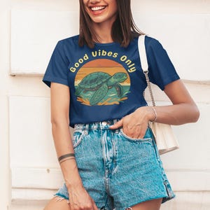 May include: Navy blue t-shirt with a turtle graphic and the text "Good Vibes Only". The turtle is green and the sun is orange and yellow. The model is wearing denim shorts.