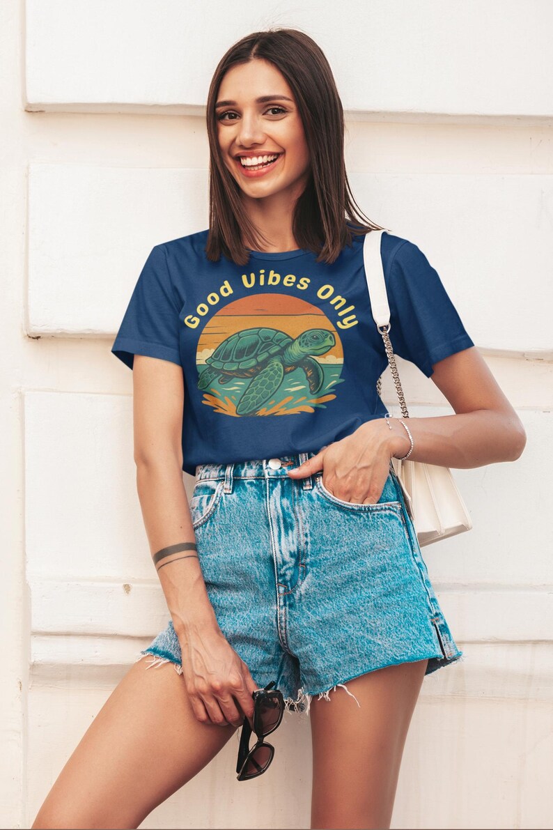 May include: Navy blue t-shirt with a turtle graphic and the text "Good Vibes Only". The turtle is green and the sun is orange and yellow. The model is wearing denim shorts.