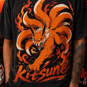 K&ouml;nnte beinhalten: Ein schwarzes T-Shirt mit kurzen &Auml;rmeln und einem Grafikdruck eines orangefarbenen neunschw&auml;nzigen Fuchses, der von stilisierten orangefarbenen und roten Flammen umgeben ist. Das Wort Kitsune ist in einer Pinselstrich-Schrift in Orange und Rot gedruckt.
