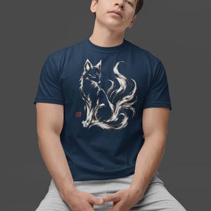 May include: A navy blue short-sleeve t-shirt featuring a white and black ink-wash style illustration of a multi-tailed fox. The graphic includes a small red square stamp mark on the left side of the fox design, set against a solid dark background.