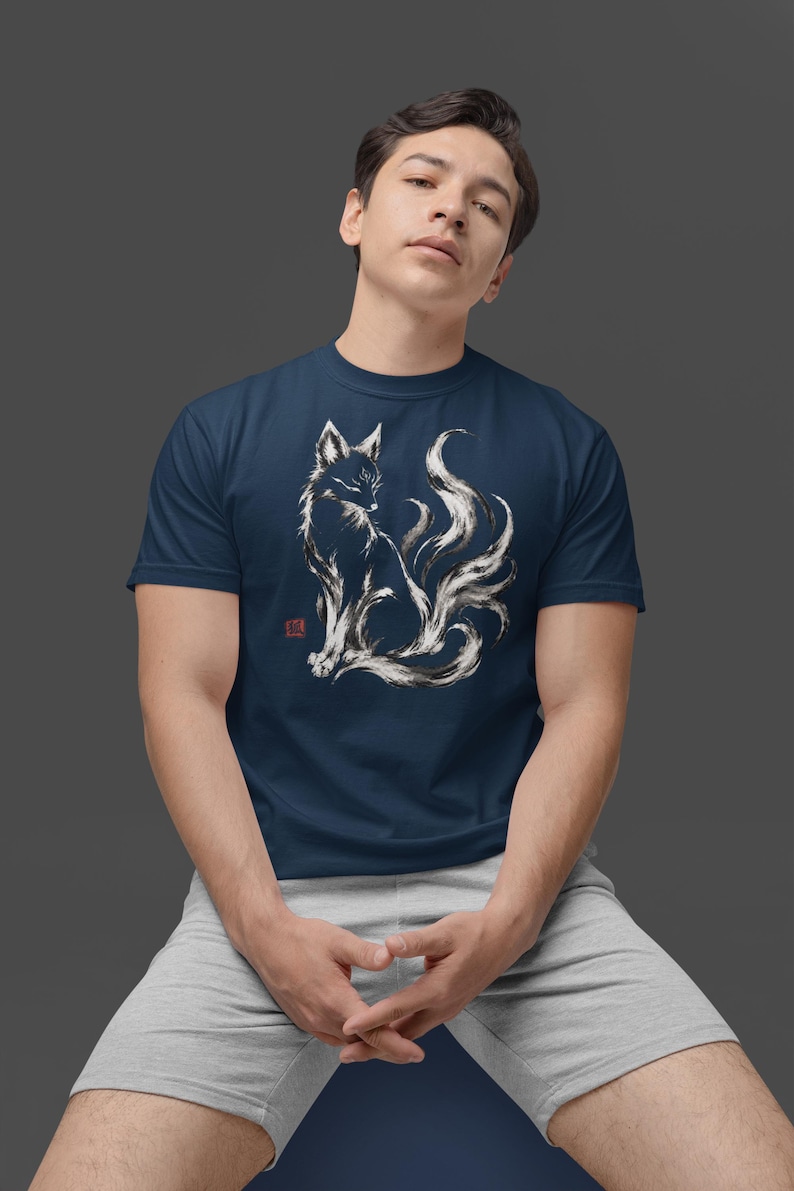 May include: A navy blue short-sleeve t-shirt featuring a white and black ink-wash style illustration of a multi-tailed fox. The graphic includes a small red square stamp mark on the left side of the fox design, set against a solid dark background.