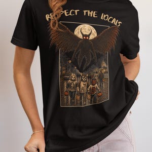 May include: A black short-sleeved t-shirt featuring a graphic print with the text "RESPECT THE LOCALS" in a tan font. The illustration depicts a large winged creature hovering above three children in costumes on a street under a full moon.