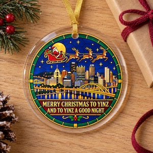 May include: A round, clear acrylic Christmas ornament featuring a stained-glass style illustration of the Pittsburgh skyline at night, Santa in a sleigh with reindeer, and the text MERRY CHRISTMAS TO YINZ AND TO YINZ A GOOD NIGHT.