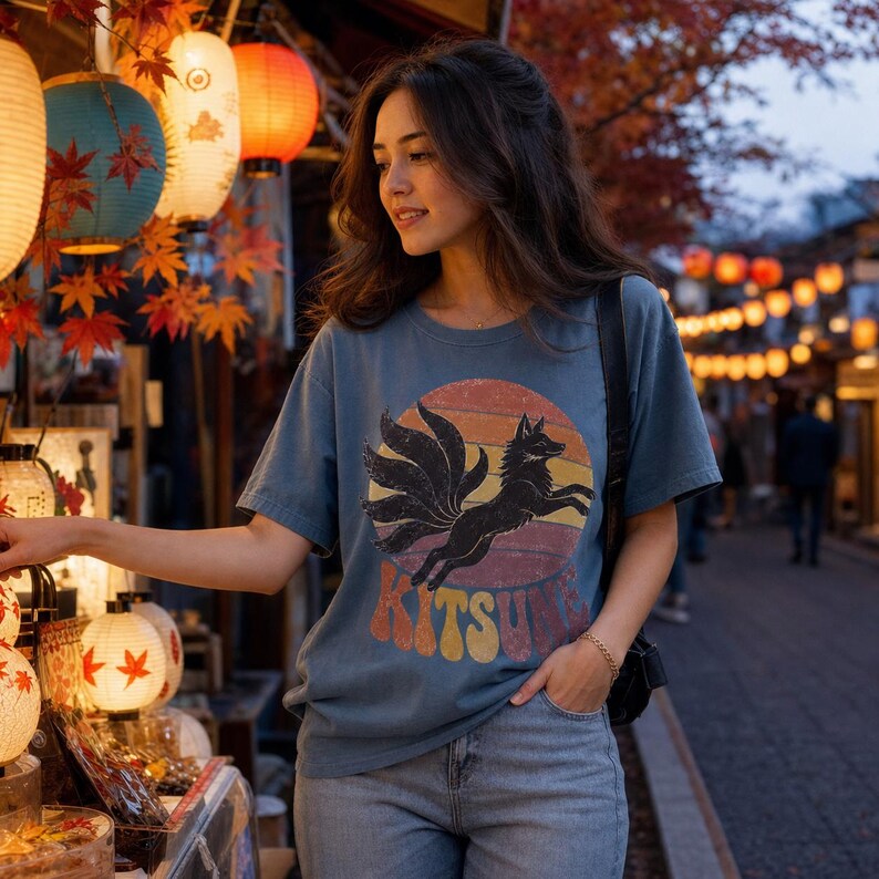 May include: A slate blue short-sleeve t-shirt featuring a graphic of a black nine-tailed fox leaping in front of a sunset-colored circle. The word KITSUNE is printed in large, retro-style block letters below the fox graphic on the front of the shirt.