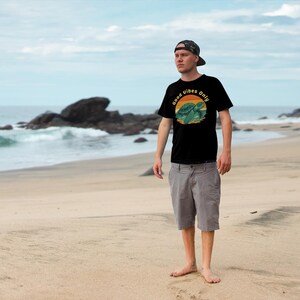 May include: Black t-shirt with a turtle graphic and the text "Good Vibes Only." The shirt is worn with grey shorts and a camouflage baseball cap. The background is a beach with ocean waves and rocks.