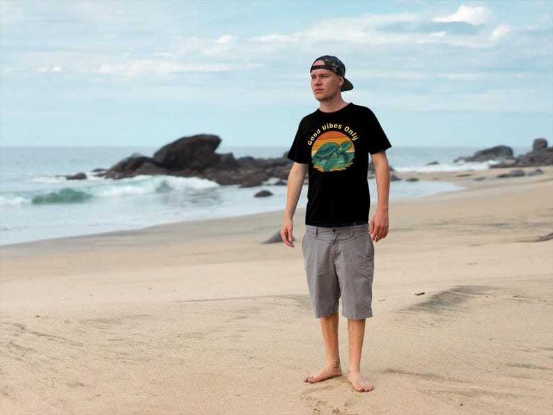 May include: Black t-shirt with a turtle graphic and the text "Good Vibes Only." The shirt is worn with grey shorts and a camouflage baseball cap. The background is a beach with ocean waves and rocks.