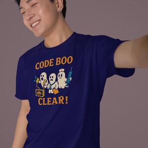 Pode incluir: Camiseta azul marinho com um gr&aacute;fico engra&ccedil;ado de tr&ecirc;s fantasmas de desenho animado, um computador e uma seringa. O texto laranja "CODE BOO CLEAR!" est&aacute; impresso. A camiseta &eacute; feita de um material macio.