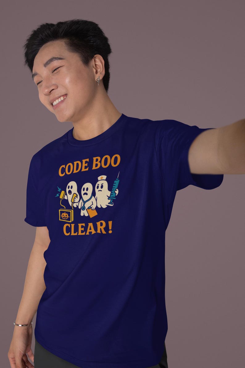 Pode incluir: Camiseta azul marinho com um gr&aacute;fico engra&ccedil;ado de tr&ecirc;s fantasmas de desenho animado, um computador e uma seringa. O texto laranja "CODE BOO CLEAR!" est&aacute; impresso. A camiseta &eacute; feita de um material macio.