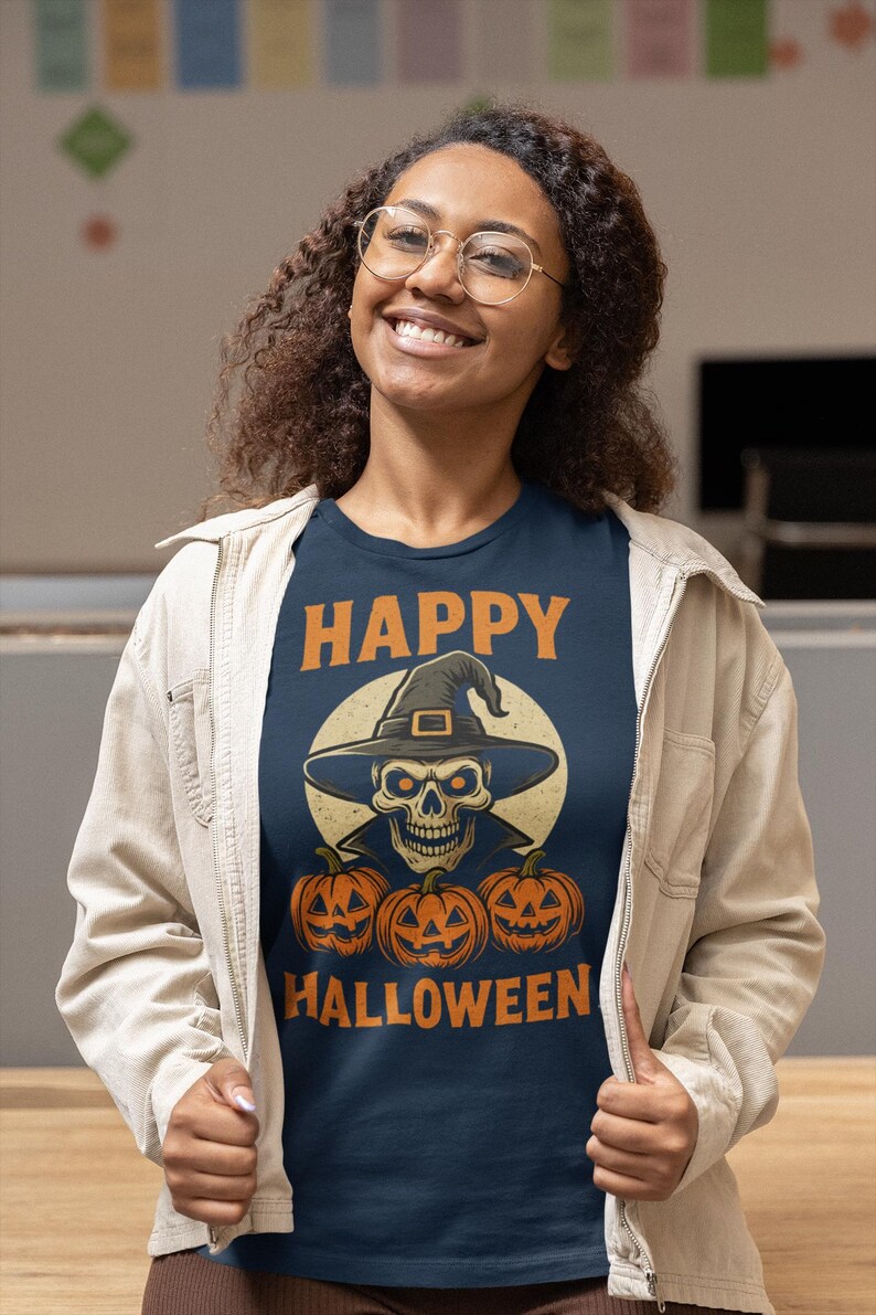 May include: Navy blue t-shirt with a Halloween design. The graphic features a skull wearing a witch hat, three carved pumpkins, and the words "HAPPY HALLOWEEN" in orange. The person is wearing a light beige jacket.