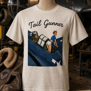 May include: A cream-colored short-sleeve t-shirt featuring the text Tail Gunner in black script above a graphic illustration of a blue military aircraft with a pilot and a person in a blue top and white shorts operating a mounted machine gun.