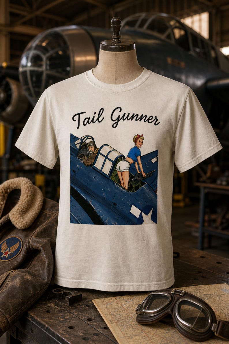 May include: A cream-colored short-sleeve t-shirt featuring the text Tail Gunner in black script above a graphic illustration of a blue military aircraft with a pilot and a person in a blue top and white shorts operating a mounted machine gun.