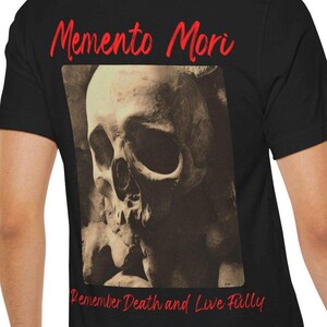 May include: Black t-shirt featuring a sepia-toned skull graphic. The shirt has the words "Memento Mori" in red script above the skull and "Remember Death and Live Fully" in red script below the skull.