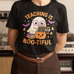 May include: Black t-shirt with a Halloween graphic. The design includes a cartoon ghost in glasses, a pumpkin, bats, and the text "TEACHING IS BOO-TIFUL" in orange. The shirt is worn with brown trousers and a brown belt.