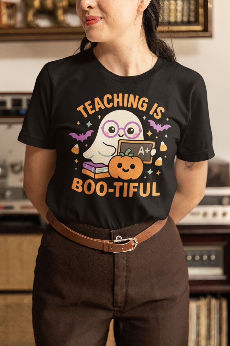 May include: Black t-shirt with a Halloween graphic. The design includes a cartoon ghost in glasses, a pumpkin, bats, and the text "TEACHING IS BOO-TIFUL" in orange. The shirt is worn with brown trousers and a brown belt.