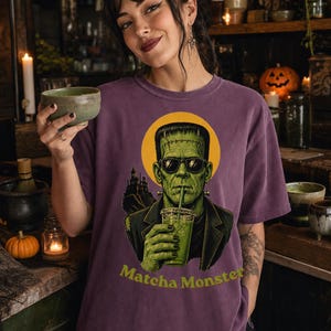 Może przedstawiać: Fioletowy t-shirt z kr&oacute;tkim rękawem z grafiką zielonego potwora w okularach przeciwsłonecznych i ciemnej marynarce, trzymającego zielony nap&oacute;j. Za potworem znajduje się ż&oacute;łte koło i sylwetka zamku. Poniżej widnieje napis Matcha Monster.