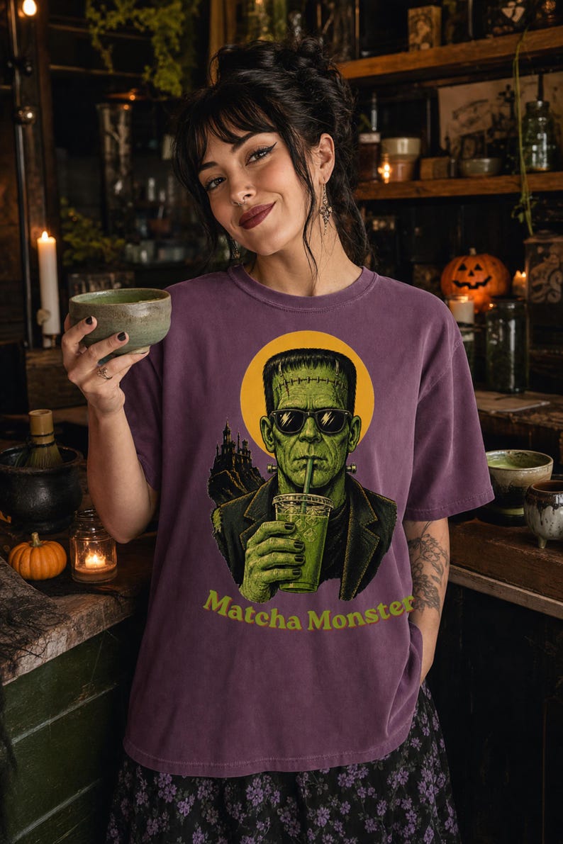 Może przedstawiać: Fioletowy t-shirt z kr&oacute;tkim rękawem z grafiką zielonego potwora w okularach przeciwsłonecznych i ciemnej marynarce, trzymającego zielony nap&oacute;j. Za potworem znajduje się ż&oacute;łte koło i sylwetka zamku. Poniżej widnieje napis Matcha Monster.