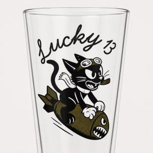 May include: A clear glass pint glass featuring a black and olive green graphic of a cartoon cat wearing goggles and a scarf, riding a bomb with shark teeth art. The text Lucky 13 is printed in a black cursive script above the cat illustration.