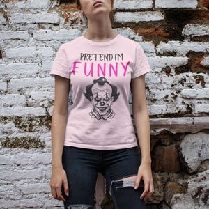 May include: A light pink t-shirt with the text "PRETEND I'M FUNNY" in black and pink. Below the text is a black and white illustration of a clown's face. The person is wearing black jeans.