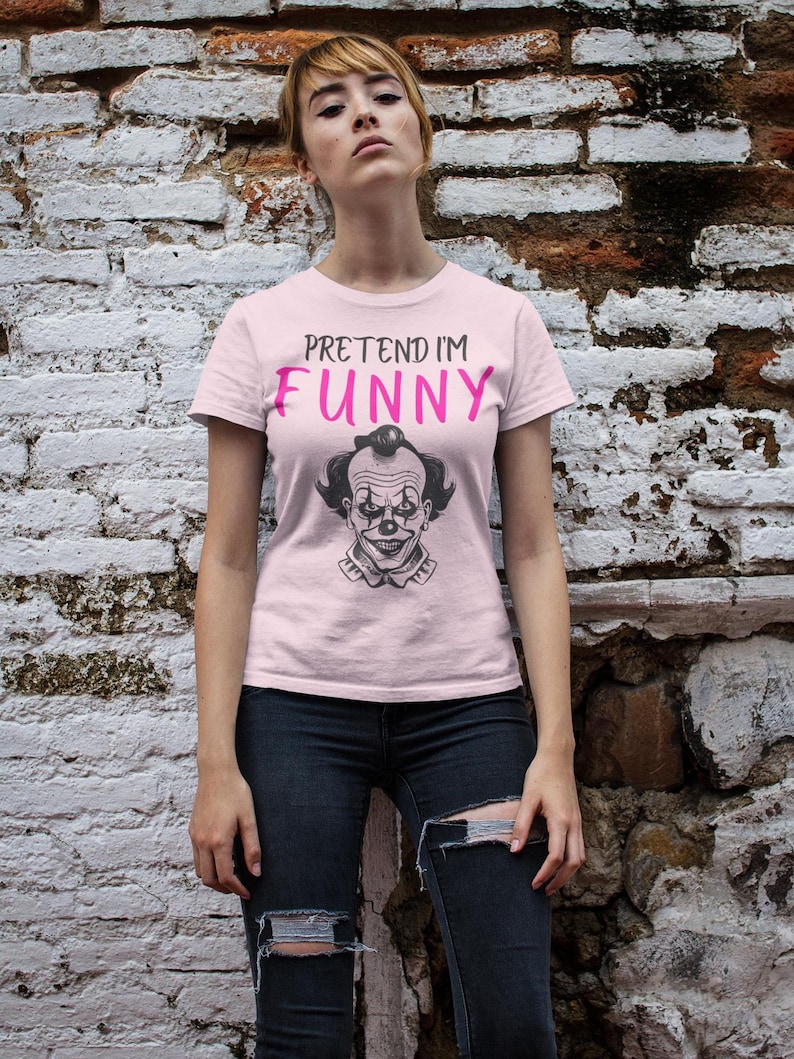 May include: A light pink t-shirt with the text "PRETEND I'M FUNNY" in black and pink. Below the text is a black and white illustration of a clown's face. The person is wearing black jeans.