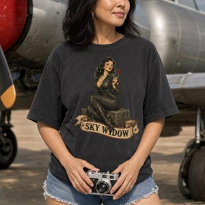 May include: A charcoal gray short-sleeve t-shirt featuring a graphic of a pin-up style figure in a black jumpsuit sitting on a crate, holding a red rose. Below the figure is a beige banner with the text SKY WYDOW and two small red stars.