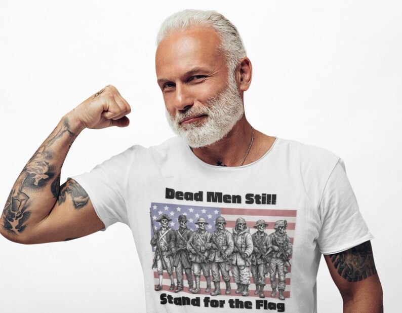 May include: A white short-sleeved t-shirt featuring a graphic print of seven skeletal soldiers in various historical military uniforms standing in front of an American flag. The text "Dead Men Still!" and "Stand for the Flag" is printed in black.
