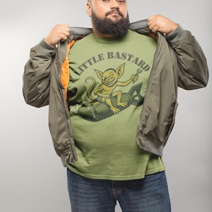 May include: Olive green bomber jacket with an orange lining, worn over a light green t-shirt. The t-shirt features a cartoon goblin riding a bomb with the text "LITTLE BASTARD". The person is wearing blue jeans.