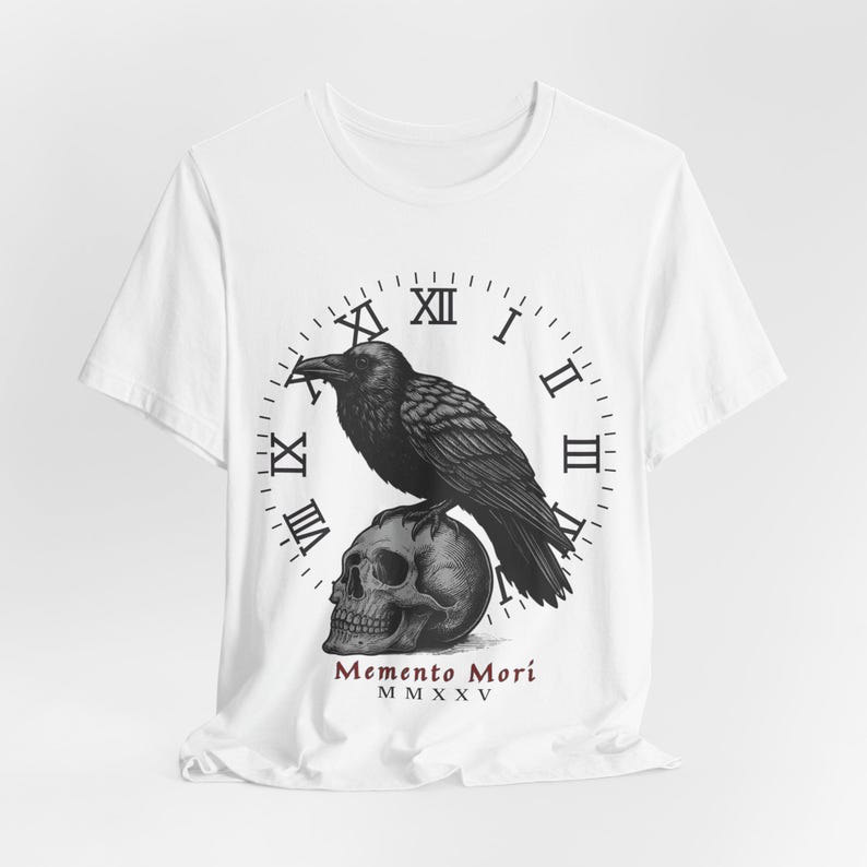 May include: White t-shirt featuring a black raven perched atop a skull, set against a clock face with Roman numerals. The phrase "Memento Mori" is printed below the skull. The design is a detailed, monochromatic illustration.