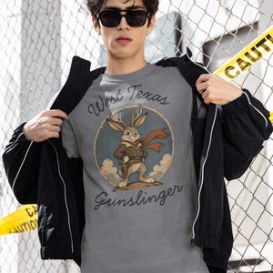 May include: A grey t-shirt featuring a circular graphic of a jackalope wearing a brown leather jacket and a red scarf, with a holster at its waist. The text West Texas Gunslinger is printed in a dark script font above and below the illustration.