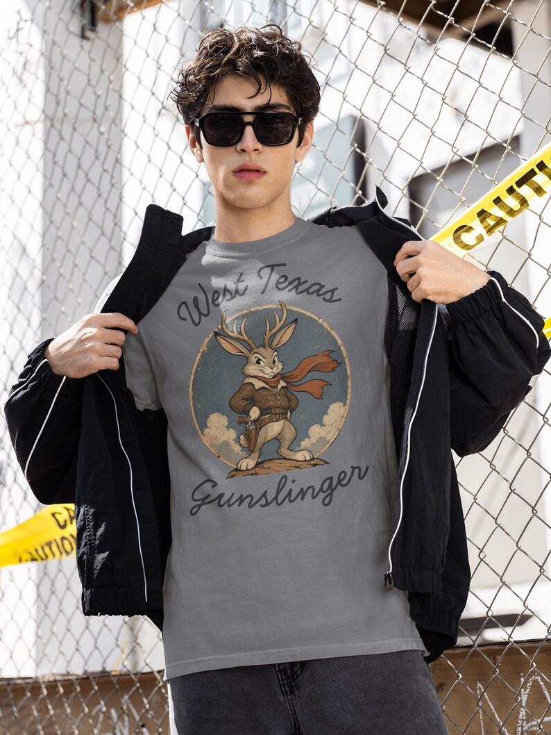 May include: A grey t-shirt featuring a circular graphic of a jackalope wearing a brown leather jacket and a red scarf, with a holster at its waist. The text West Texas Gunslinger is printed in a dark script font above and below the illustration.
