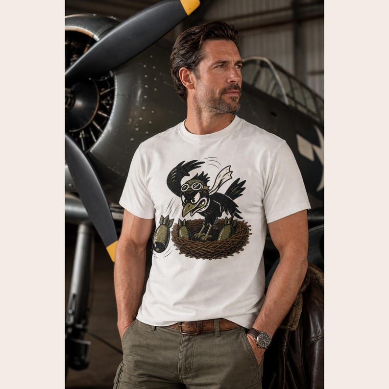 Puede incluir: Una camiseta blanca de manga corta con una ilustraci&oacute;n gr&aacute;fica de un p&aacute;jaro negro con gafas de aviador, posado en un nido y lanzando bombas de color verde oliva. El p&aacute;jaro lleva una bufanda blanca y tiene una expresi&oacute;n agresiva de dibujos.