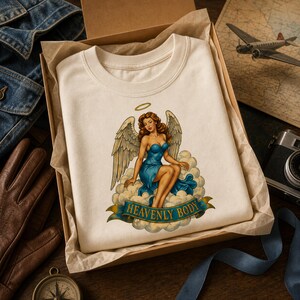 May include: A cream-colored t-shirt featuring a graphic of an angel with brown hair, large wings, and a blue dress sitting on clouds. Below the angel, a blue banner contains the text HEAVENLY BODY in gold lettering. The shirt is inside a box.