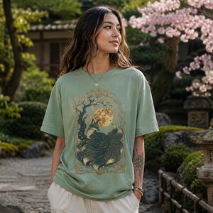 May include: A sage green short-sleeved t-shirt featuring a detailed graphic print of a multi-tailed fox sitting beneath a large full moon and a flowering tree, framed by an ornate gold-coloured border with intricate swirling patterns and foliage.