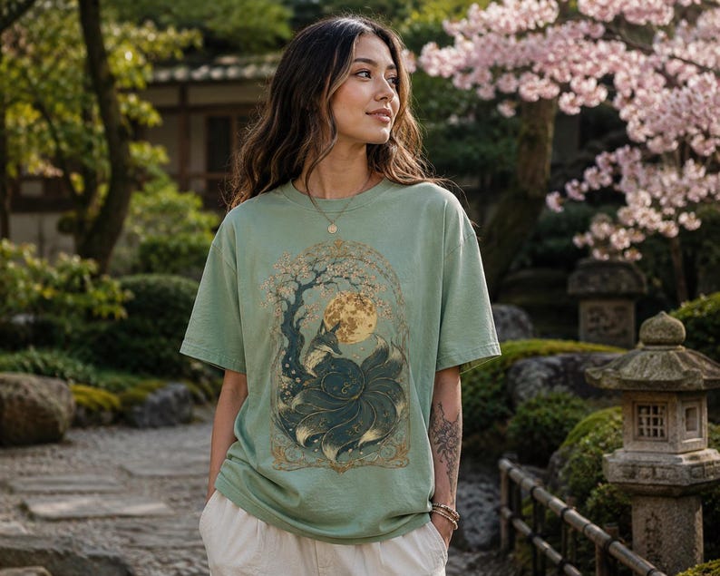 May include: A sage green short-sleeved t-shirt featuring a detailed graphic print of a multi-tailed fox sitting beneath a large full moon and a flowering tree, framed by an ornate gold-coloured border with intricate swirling patterns and foliage.