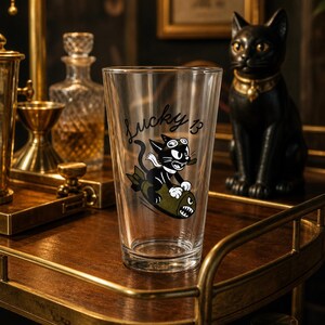 May include: A clear glass pint tumbler featuring a black and white cartoon cat wearing goggles and riding an olive green bomb. The text "Lucky 13" is printed in black script above the cat illustration. The glass has a tapered cylindrical shape.