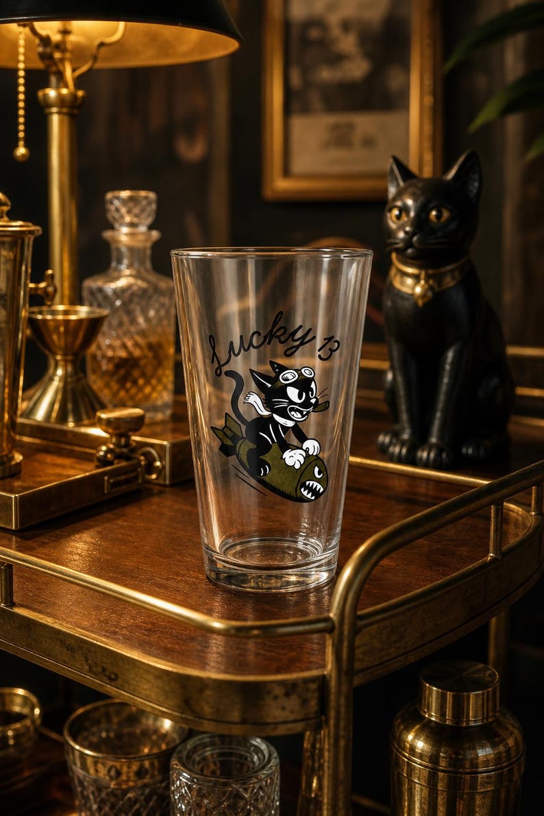 May include: A clear glass pint tumbler featuring a black and white cartoon cat wearing goggles and riding an olive green bomb. The text "Lucky 13" is printed in black script above the cat illustration. The glass has a tapered cylindrical shape.