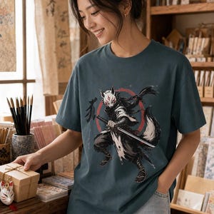May include: A dark teal short-sleeve t-shirt featuring a graphic print of a stylized fox warrior wearing a white mask and holding a katana sword. The design includes a red circular background element and black Japanese calligraphy characters.