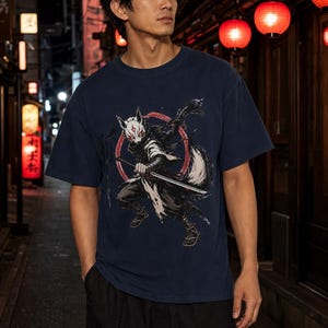 May include: A navy blue short-sleeve t-shirt featuring a graphic print of a masked figure with fox-like features holding a katana sword. The design includes a red circular element behind the figure and stylized black brushstroke accents.