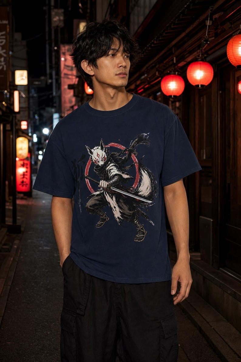 May include: A navy blue short-sleeve t-shirt featuring a graphic print of a masked figure with fox-like features holding a katana sword. The design includes a red circular element behind the figure and stylized black brushstroke accents.