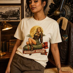 Peut inclure: Un t-shirt &agrave; manches courtes blanc cass&eacute; avec une impression graphique repr&eacute;sentant un personnage pin-up blond en veste en cuir marron assis sur une bombe verte. Le texte MIDNIGHT MISSION est imprim&eacute; sur une banni&egrave;re rouge en bas du motif.