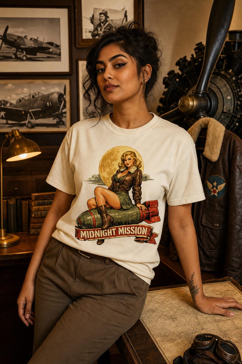 Peut inclure: Un t-shirt &agrave; manches courtes blanc cass&eacute; avec une impression graphique repr&eacute;sentant un personnage pin-up blond en veste en cuir marron assis sur une bombe verte. Le texte MIDNIGHT MISSION est imprim&eacute; sur une banni&egrave;re rouge en bas du motif.
